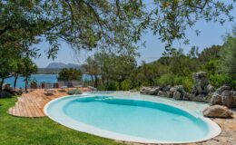 Serene outdoor swimming pool surrounded by lush greenery and natural rock formations, with a picturesque view of the water and mountains in the background. Ideal for relaxation and leisure.