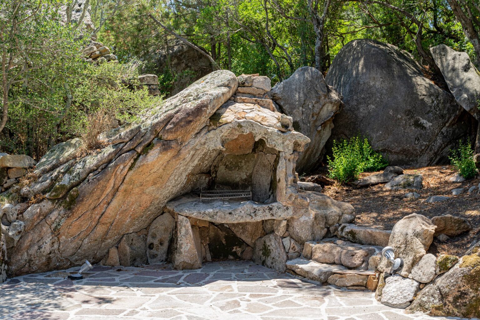 Natural stone shelter surrounded by greenery and large rocks, featuring a flat stone bench and steps leading to a patio area. Ideal for outdoor relaxation and nature appreciation. | Only on SopranoVillas