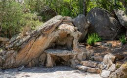 Natural stone shelter surrounded by greenery and large rocks, featuring a flat stone bench and steps leading to a patio area. Ideal for outdoor relaxation and nature appreciation.