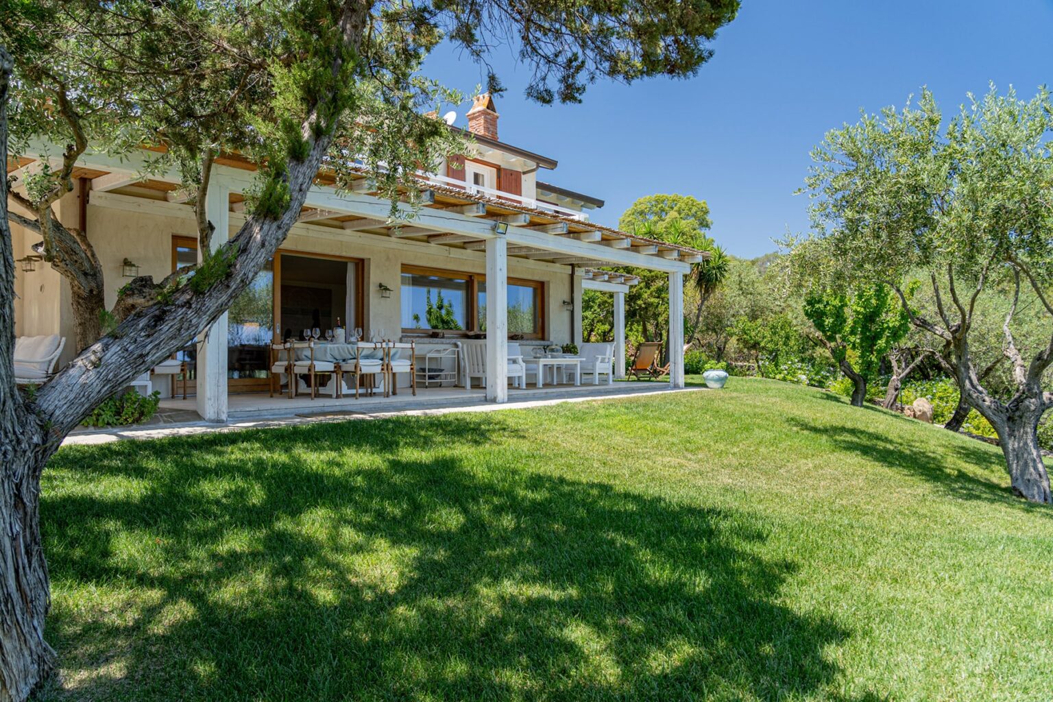 Beautiful villa with a spacious porch overlooking a lush green lawn, surrounded by olive trees under a clear blue sky. Ideal for outdoor relaxation and entertaining. | Only on SopranoVillas