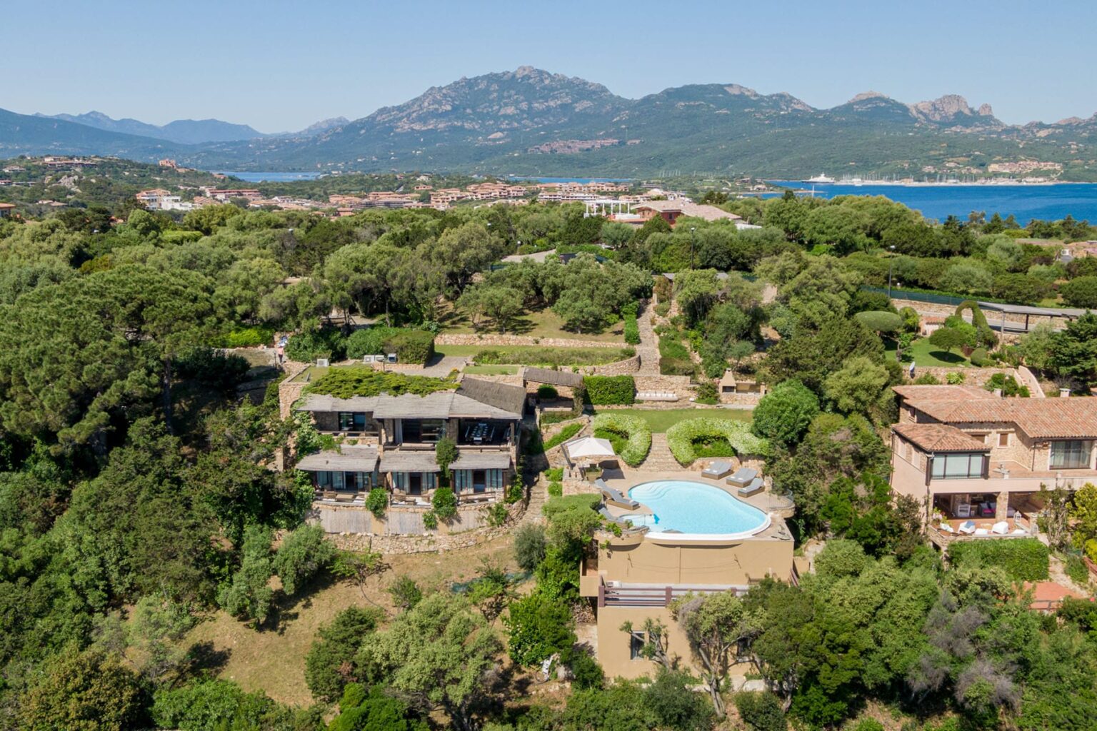 Aerial view of a luxurious villa surrounded by lush greenery, featuring a swimming pool and scenic mountains in the background, located near the coast. Ideal for vacation rentals or real estate listings. | Only on SopranoVillas