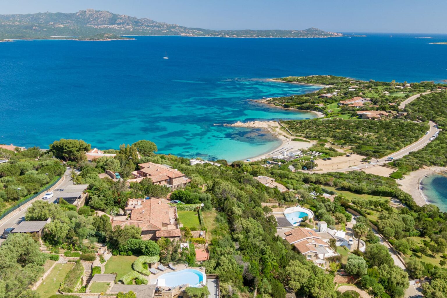 Aerial view of a scenic coastal landscape featuring lush greenery, luxurious villas, and clear turquoise waters along a serene bay. | Only on SopranoVillas