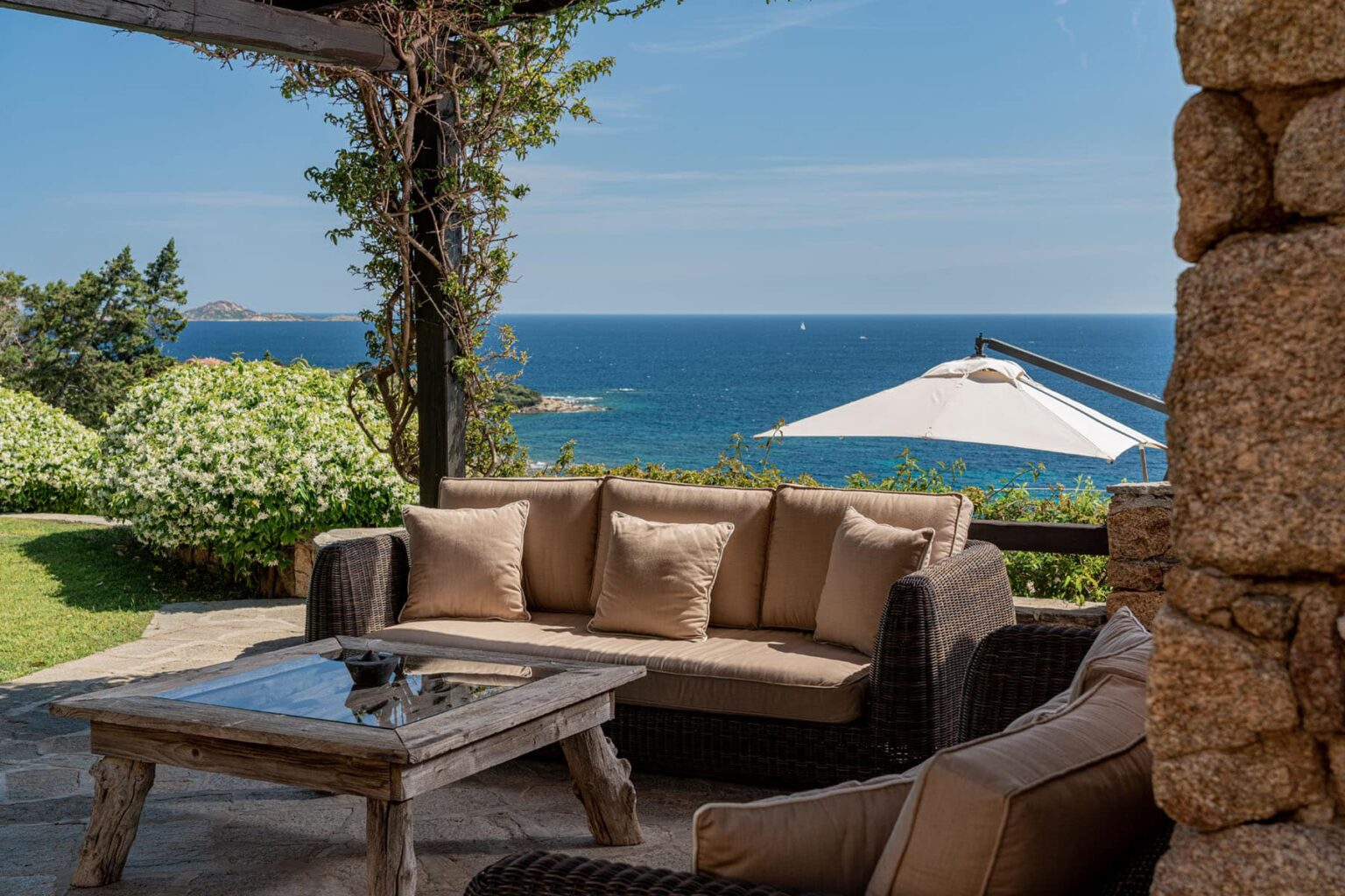 Cozy outdoor seating area with beige cushions, a wooden table, and a view of the ocean under a clear blue sky, surrounded by lush greenery and a sunshade. Ideal for relaxation and enjoying coastal scenery. | Only on SopranoVillas