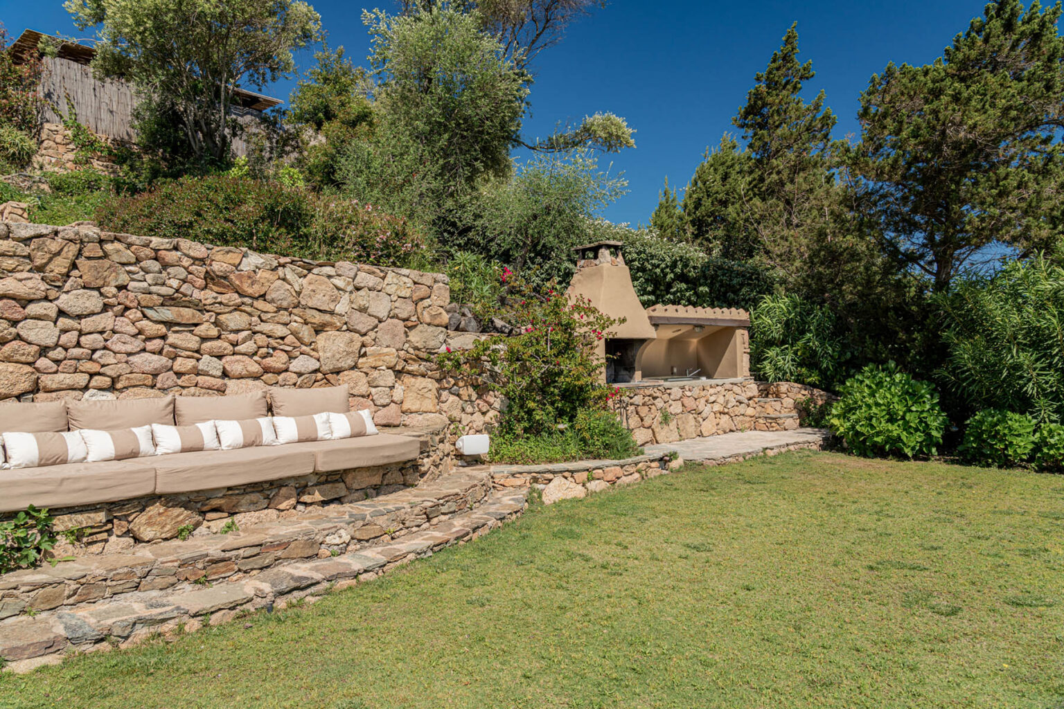 Outdoor living space featuring a stone wall, comfortable seating area with cushions, and a built-in fireplace, surrounded by lush greenery and blue skies. Ideal for relaxation and gatherings. | Only on SopranoVillas