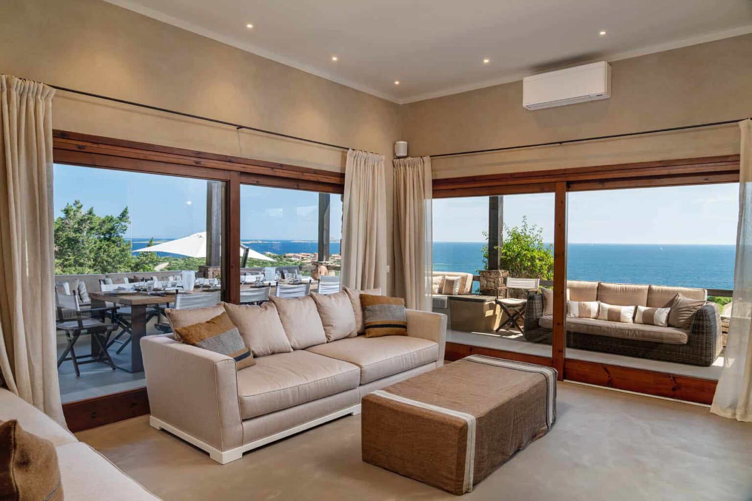 Modern living room with large windows offering ocean views, featuring a comfortable beige sofa, an ottoman, and elegant curtains. Bright and airy ambiance with natural light highlighting the stylish interior design. | Only on SopranoVillas