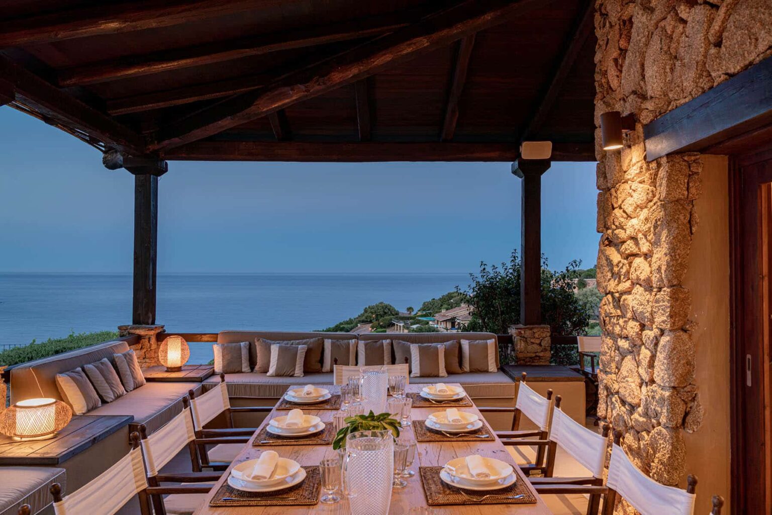Cozy outdoor dining area with a long wooden table set for a meal, surrounded by comfortable seating and illuminated by soft lighting, overlooking a serene ocean view at dusk. | Only on SopranoVillas