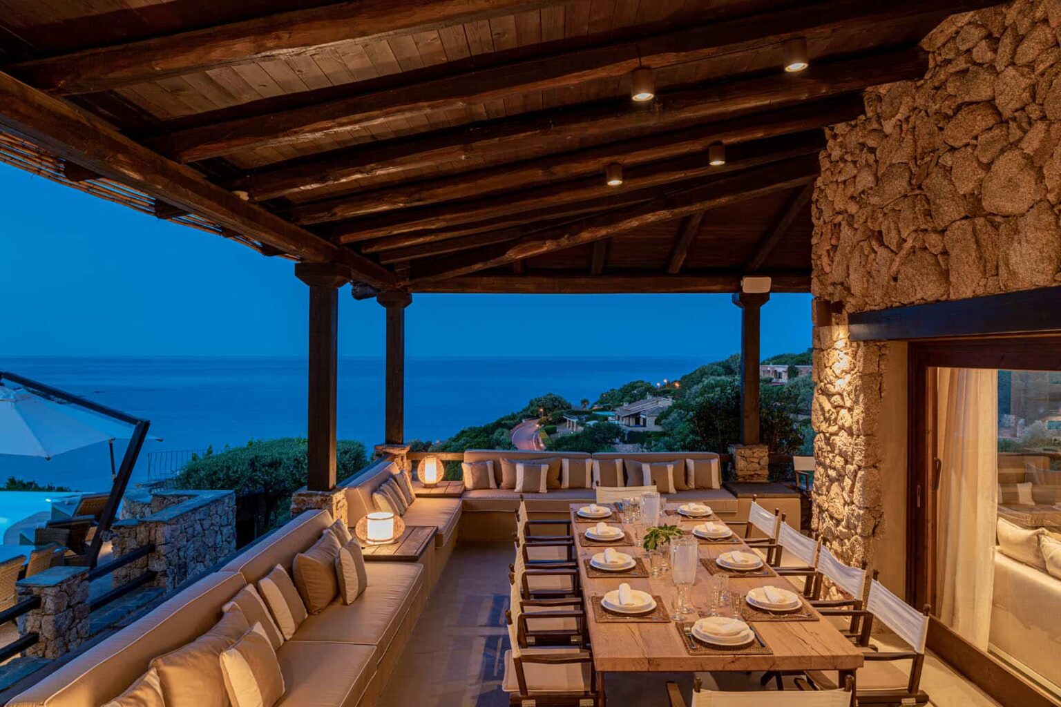 Cozy outdoor dining area with a wooden roof, stone accents, and a stunning ocean view at dusk, featuring a large table set for a meal. | Only on SopranoVillas
