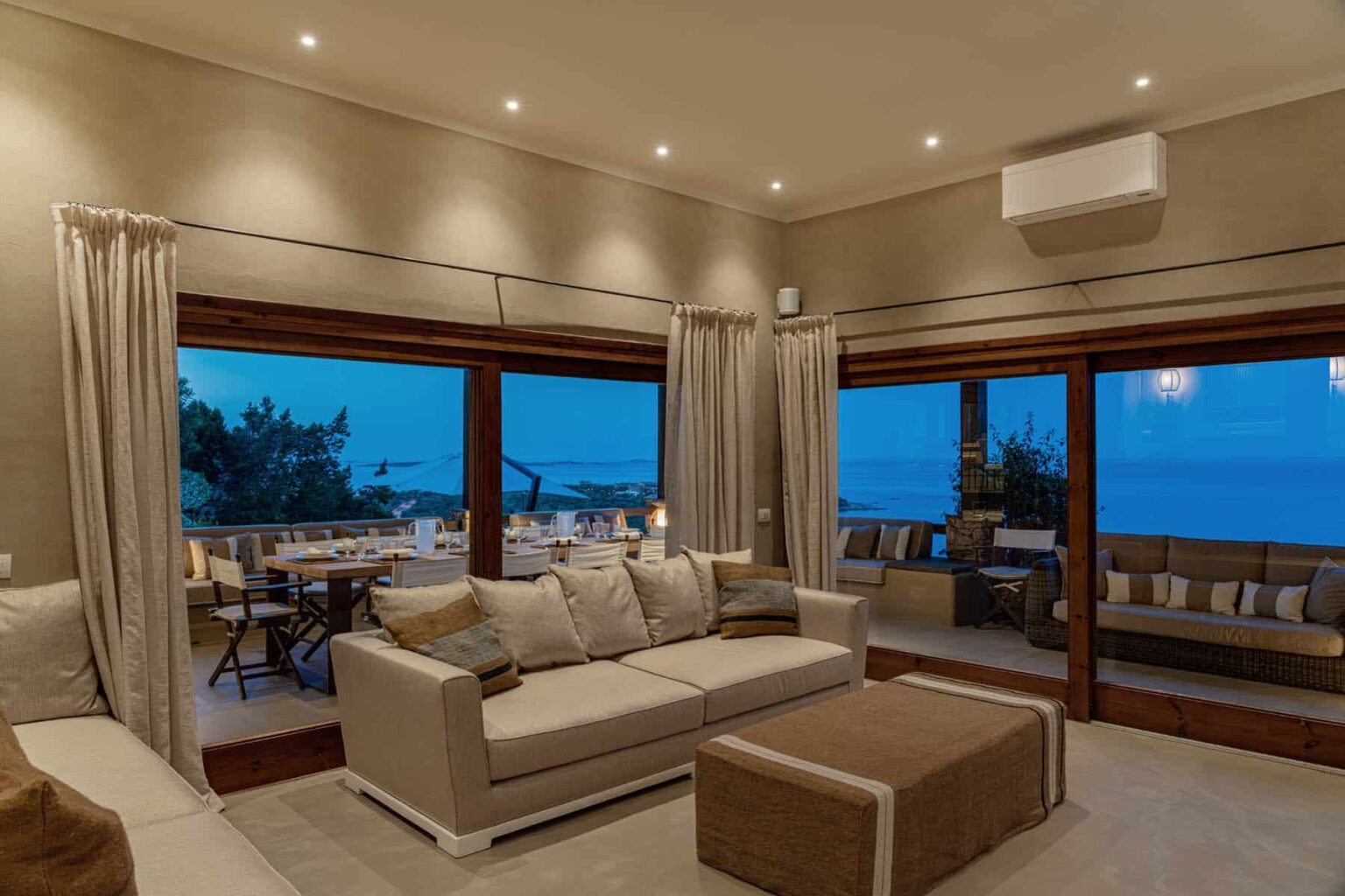 Modern living room with large windows showcasing a scenic view, featuring a comfortable beige sofa, stylish decor, and warm lighting. Ideal for relaxation and entertaining guests. | Only on SopranoVillas