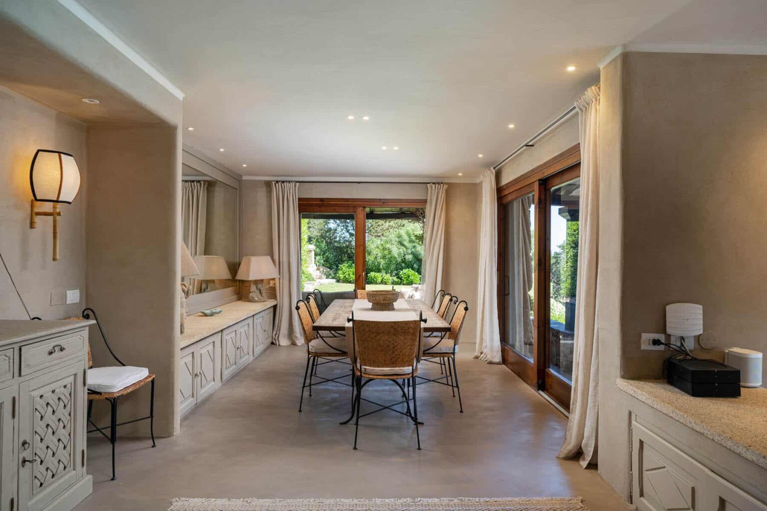 Modern dining room with a large wooden table surrounded by stylish chairs, natural light streaming through large windows, and neutral-colored walls creating a warm atmosphere. | Only on SopranoVillas