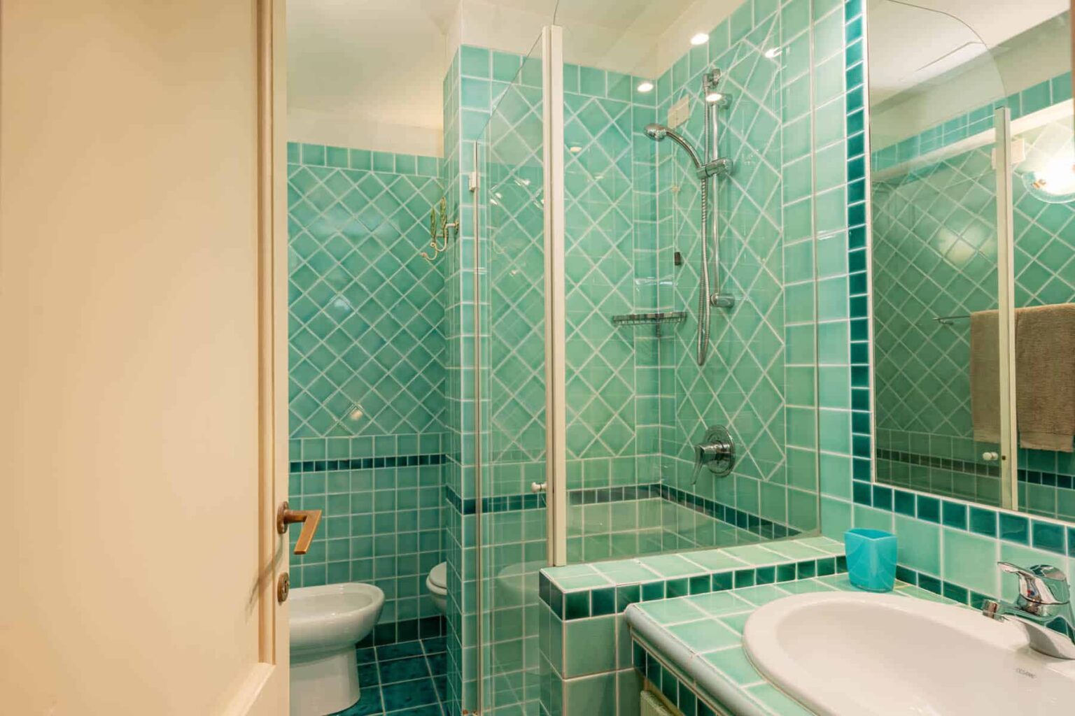 Modern bathroom featuring turquoise tiled walls, a glass shower enclosure, a white bathtub, and a toilet, creating a vibrant and stylish space. | Only on SopranoVillas
