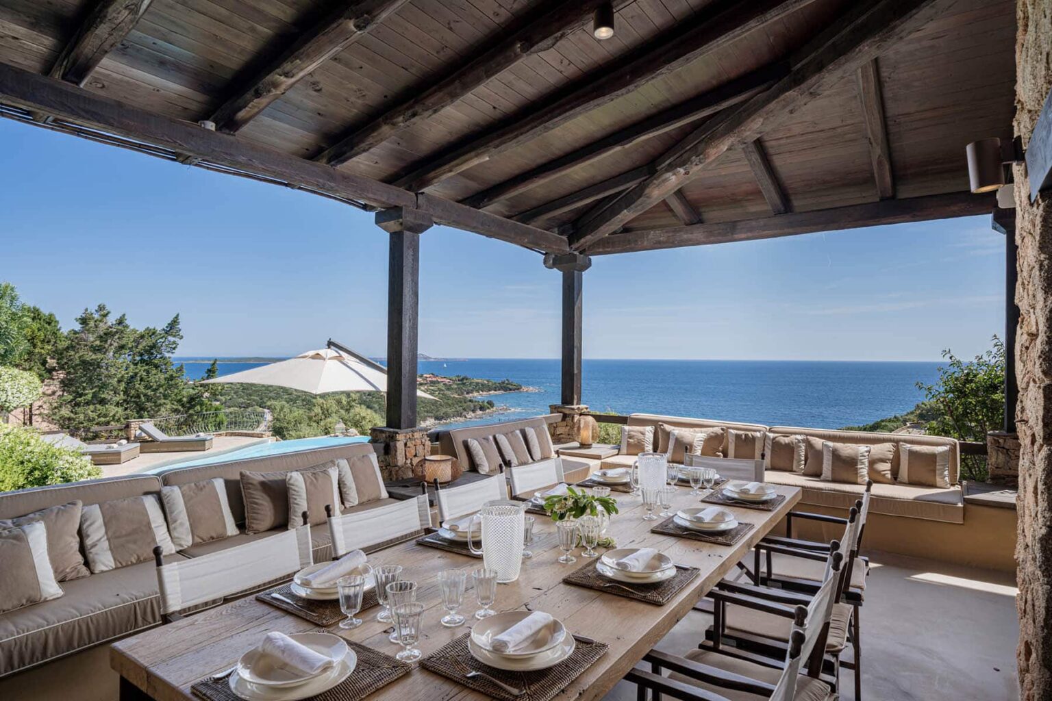 Outdoor dining area with a large wooden table set for a meal, featuring elegant tableware and comfortable seating, overlooking a scenic ocean view under a wooden pergola. Ideal for alfresco dining and entertaining guests. | Only on SopranoVillas