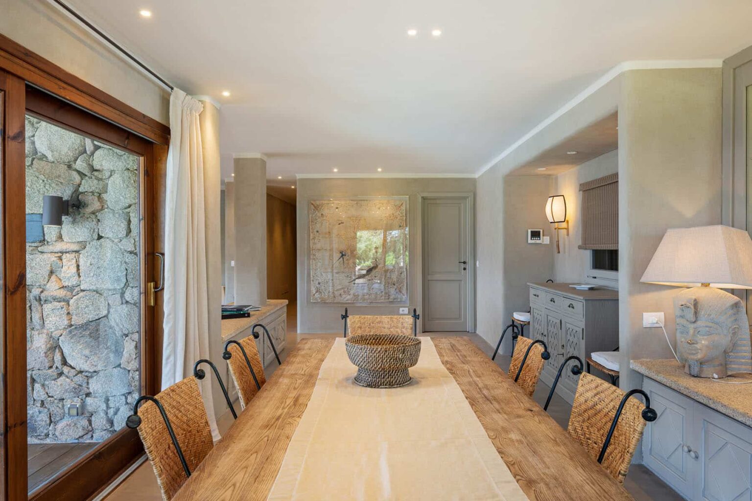 Spacious modern dining room featuring a long wooden table, stylish woven chairs, and natural light from large windows, complemented by elegant decor and a stone wall. | Only on SopranoVillas