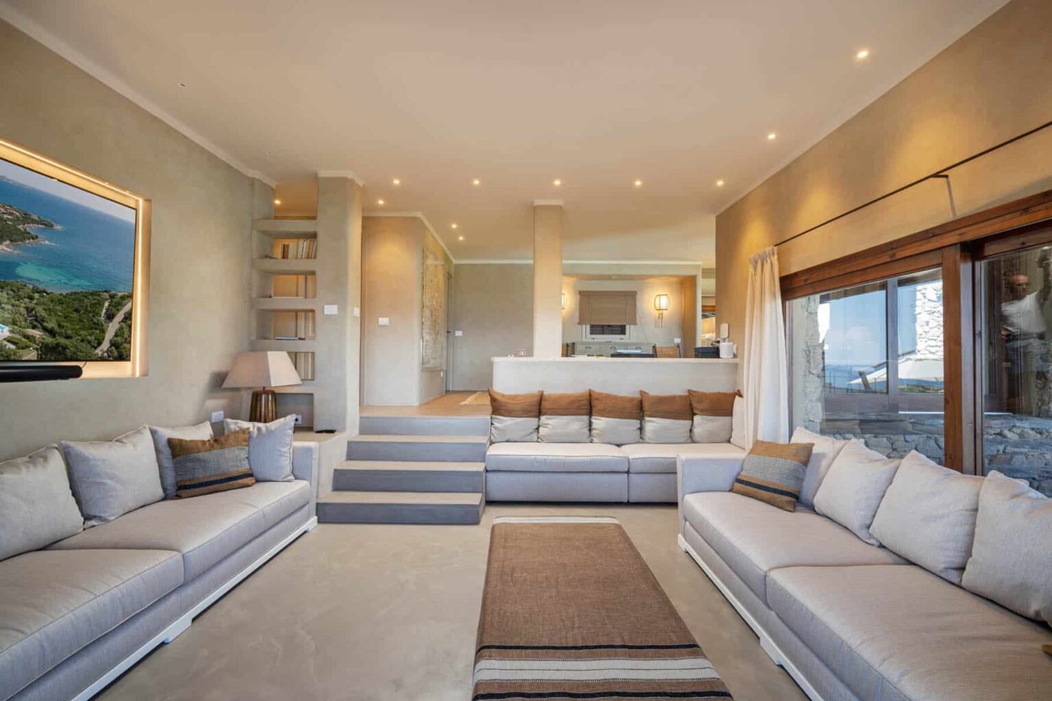 Modern living room with spacious gray sectional sofas, natural light from large windows, and a stylish decor featuring a wall-mounted TV and artwork. The open layout leads to a kitchen area, creating a comfortable and inviting atmosphere. | Only on SopranoVillas