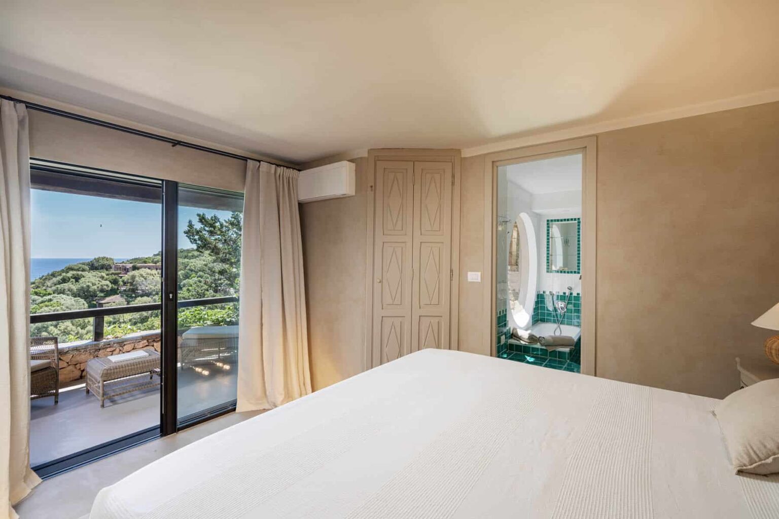 Cozy bedroom with a large bed, featuring a sliding glass door that opens to a balcony with scenic views, and a stylish en-suite bathroom visible through a mirror. The room is decorated in neutral tones with natural light streaming in. | Only on SopranoVillas