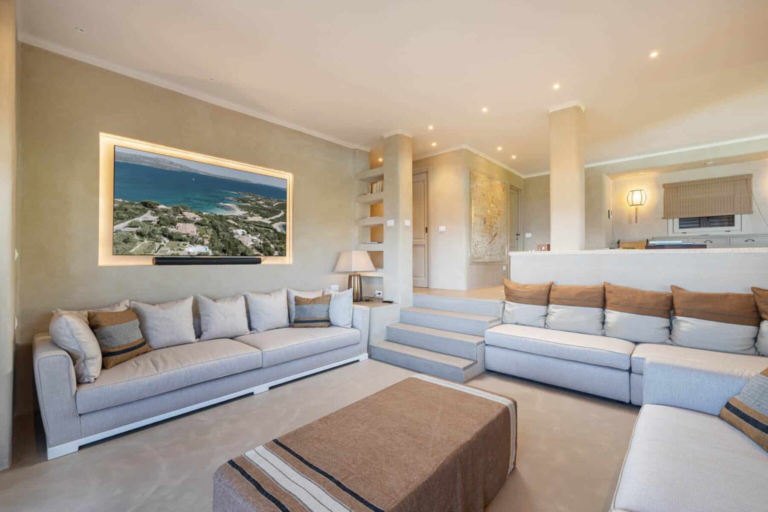 Modern living room featuring a spacious gray sectional sofa with decorative pillows, a stylish coffee table, and an illuminated wall-mounted artwork showcasing a scenic view. The open layout connects to a kitchen area with soft lighting, enhancing the contemporary aesthetic. | Only on SopranoVillas