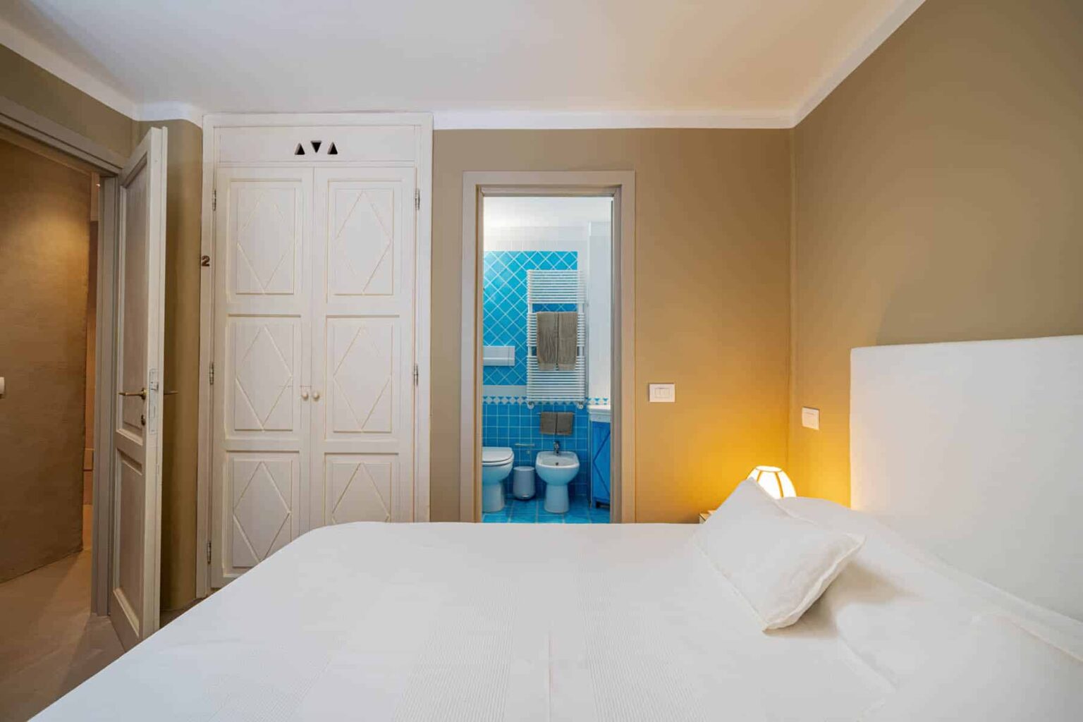 Cozy bedroom featuring a white bed with soft linens, leading to a brightly colored bathroom with blue tiles. The room has warm beige walls and a modern aesthetic, creating a welcoming atmosphere. | Only on SopranoVillas