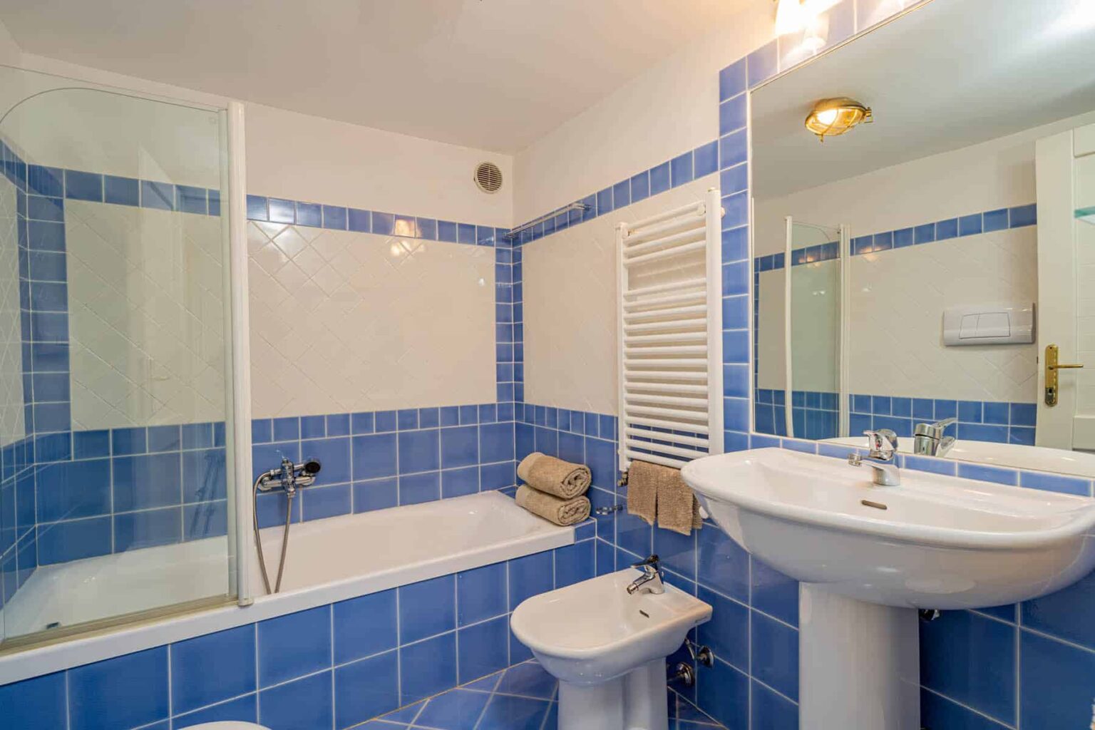 Modern bathroom featuring blue tiled walls, a bathtub with a shower, a pedestal sink, and a bidet, along with neatly arranged towels. | Only on SopranoVillas