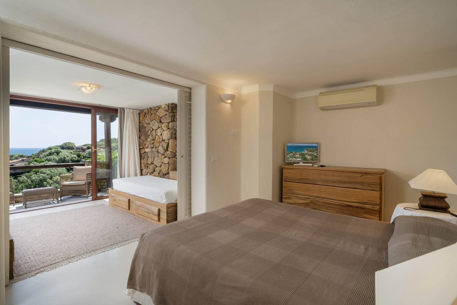 Cozy hotel room featuring a comfortable bed, wooden dresser, and a view of a balcony with outdoor seating and scenic greenery. Ideal for relaxation and enjoying nature. | Only on SopranoVillas