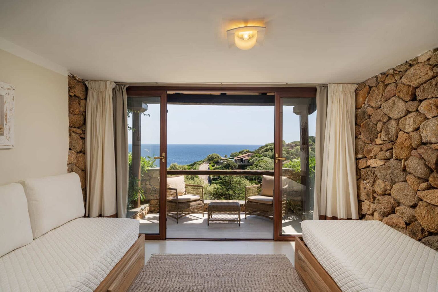 Cozy interior of a modern room featuring two single beds, a stone wall, and large glass doors opening to a scenic ocean view with a balcony. | Only on SopranoVillas