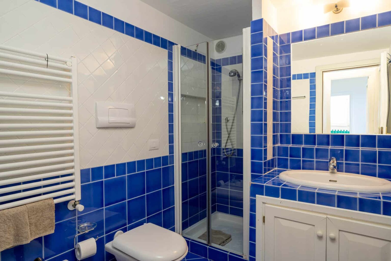 Modern bathroom featuring blue tiled walls, a glass shower enclosure, a white vanity with a sink, and a towel warmer. Ideal for showcasing contemporary bathroom design and decor. | Only on SopranoVillas