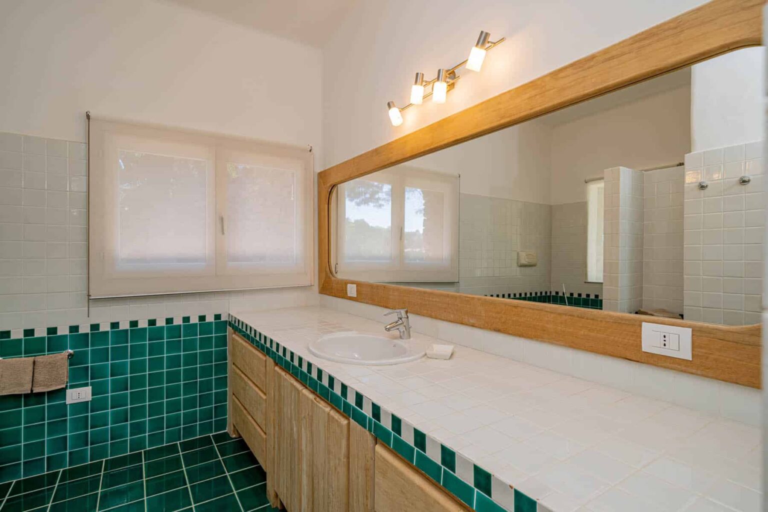 Modern bathroom interior featuring a spacious vanity with a large mirror, wooden cabinetry, and green tiled flooring. Natural light filters through a window, enhancing the bright and airy atmosphere. | Only on SopranoVillas