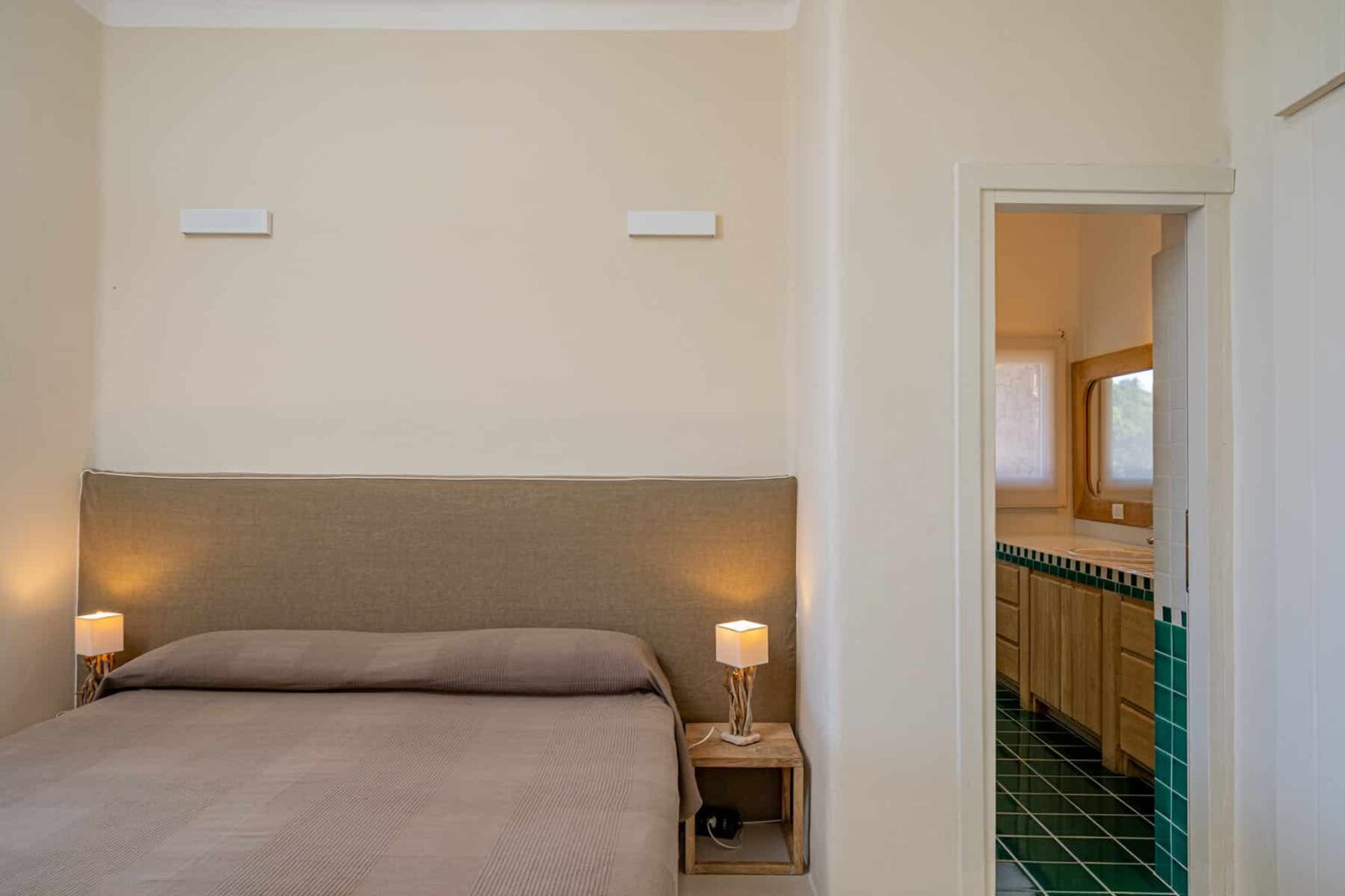 Cozy bedroom featuring a soft bed with a neutral-colored headboard, bedside lamps, and an open doorway leading to a stylish bathroom with wooden cabinetry and green tiled flooring. | Only on SopranoVillas