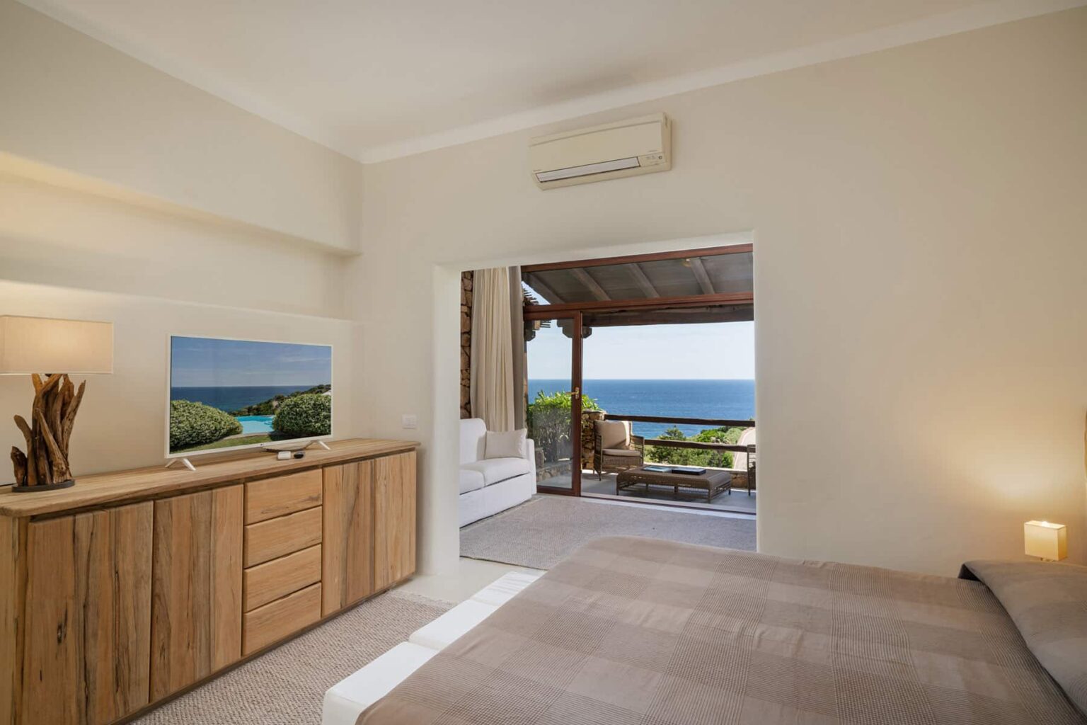 Modern bedroom with wooden dresser and flat-screen TV, featuring a large open doorway that leads to a balcony with ocean views. Bright, airy atmosphere with minimalist decor and natural light. | Only on SopranoVillas