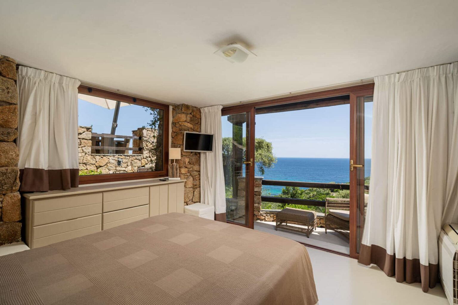 Modern bedroom with a stunning ocean view, featuring a comfortable bed, large windows, and natural stone accents. The room is bright and airy, with light curtains and a minimalist design. | Only on SopranoVillas