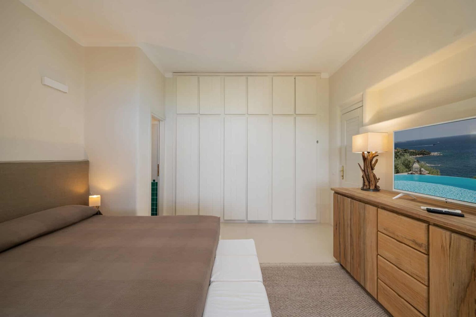 Modern bedroom interior featuring a cozy bed with brown linens, minimalist design, wooden furniture, and a wall-mounted television. The space includes ample storage with built-in wardrobes and a warm ambiance created by soft lighting. | Only on SopranoVillas
