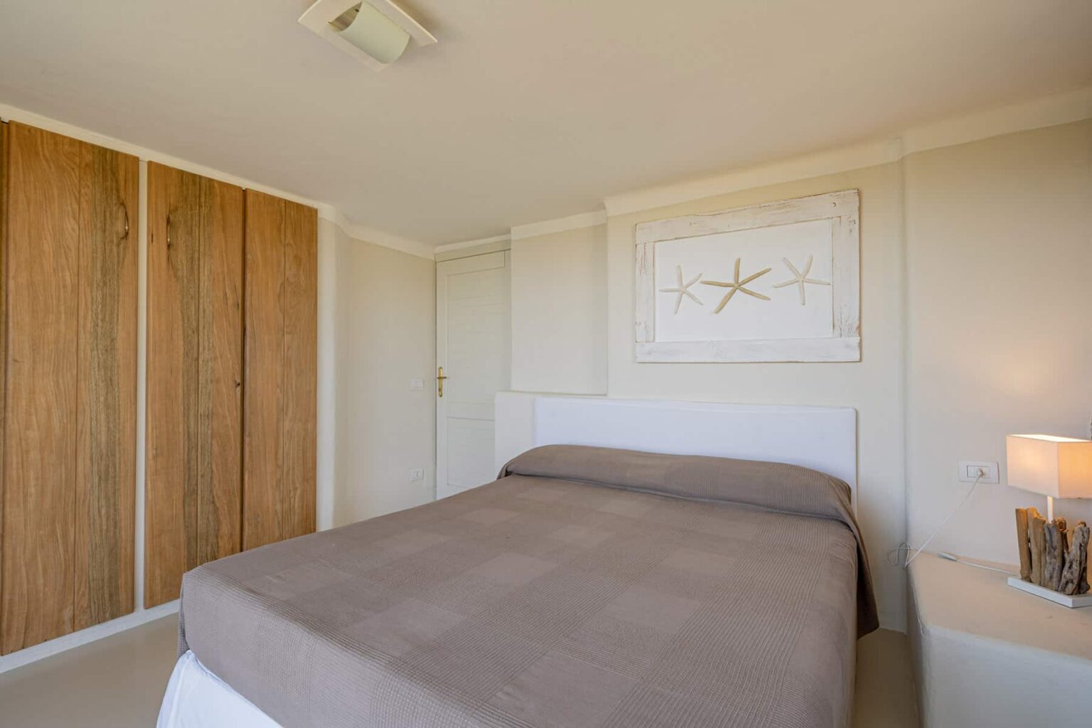 Cozy bedroom featuring a modern design with a queen-sized bed, neutral-toned bedding, wooden closet doors, and minimalist decor. A bedside lamp adds warmth to the inviting atmosphere. | Only on SopranoVillas
