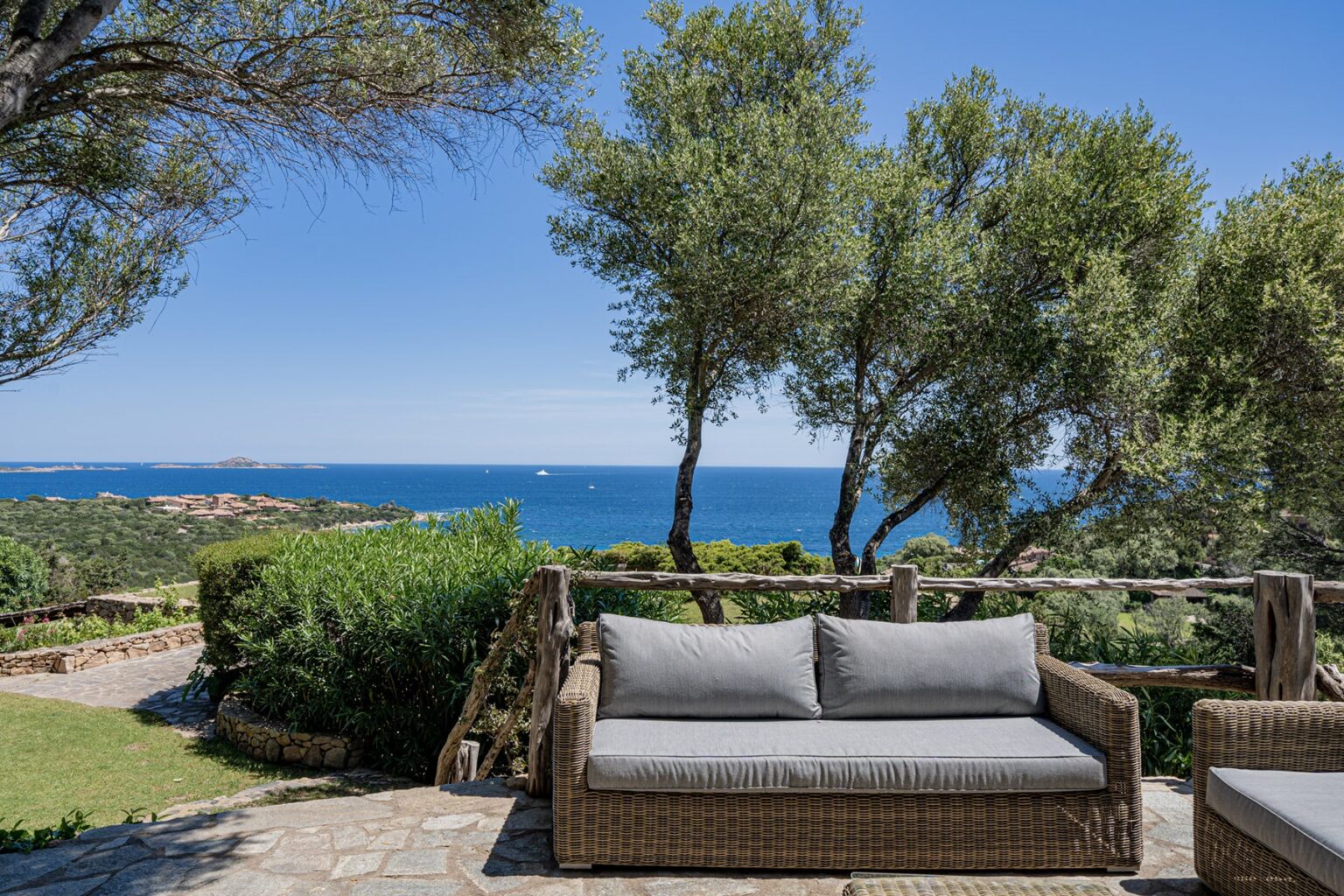 Cozy outdoor seating area with a wicker sofa overlooking a scenic view of the ocean and lush greenery under a clear blue sky. Ideal for relaxation and enjoying nature. | Only on SopranoVillas