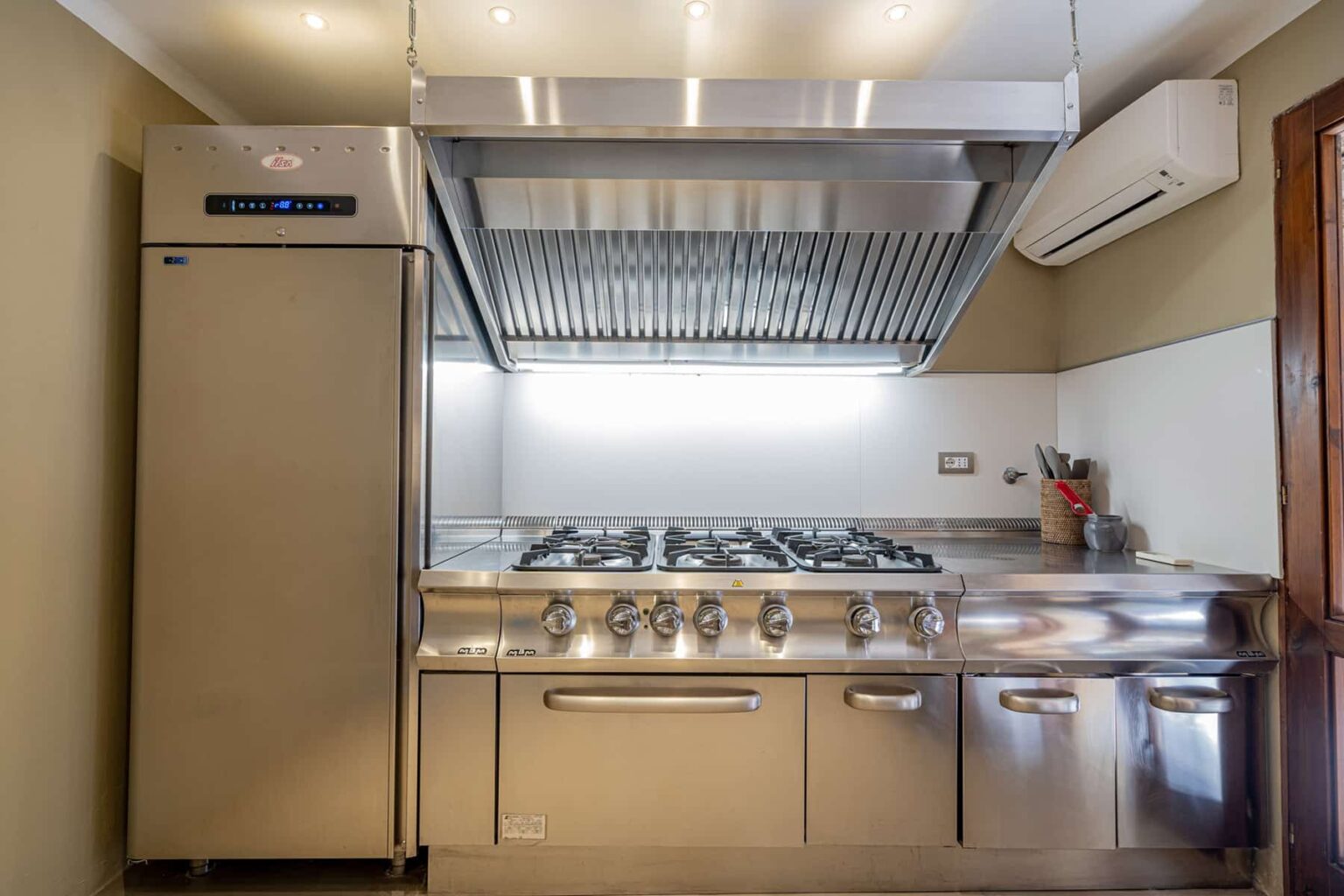 Modern kitchen featuring stainless steel appliances, including a large refrigerator and a gas range with multiple burners, complemented by a sleek exhaust hood and an air conditioning unit. | Only on SopranoVillas