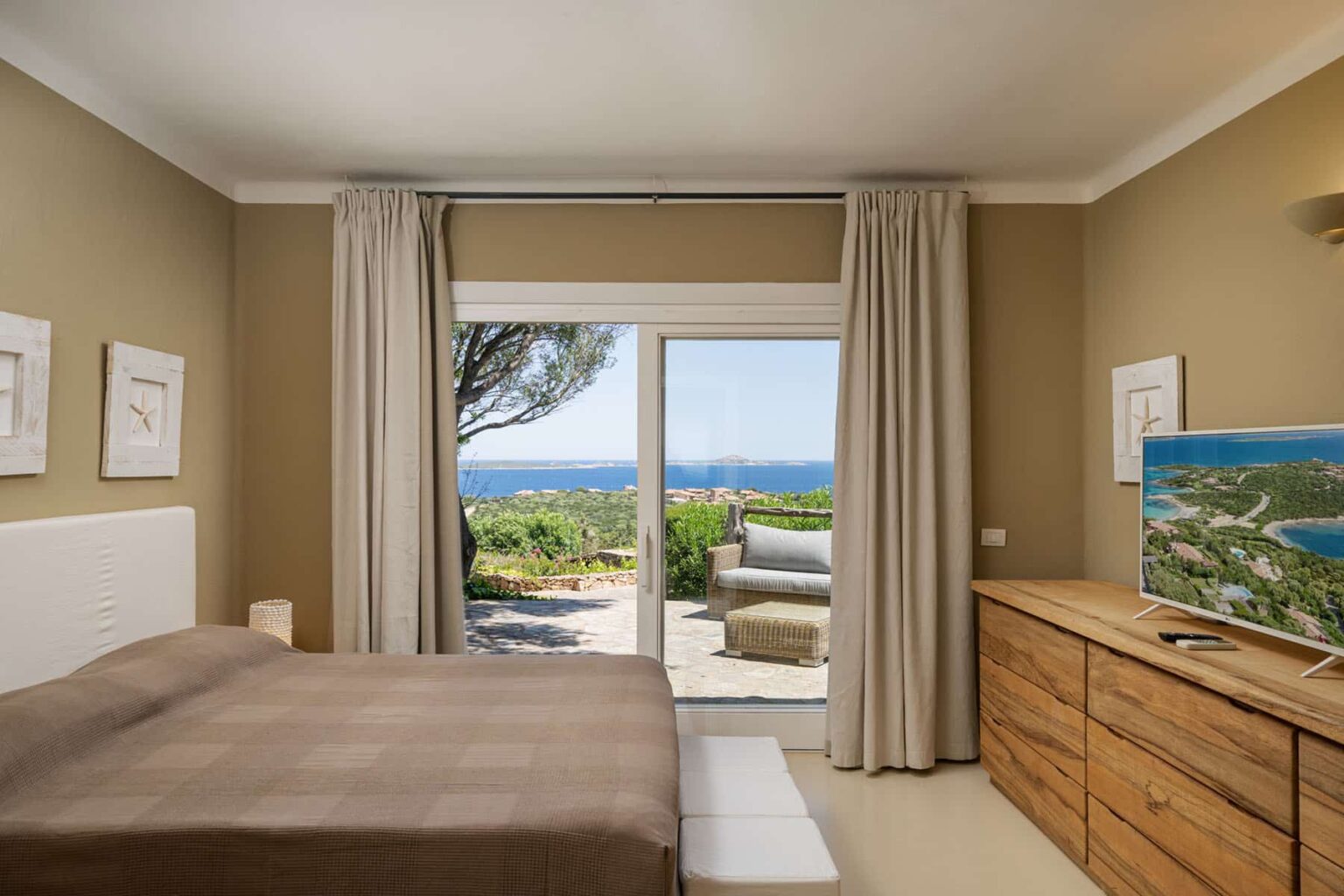 Cozy bedroom with a large window showcasing a stunning ocean view, featuring a queen-sized bed with neutral bedding, wooden dresser, and light beige walls. | Only on SopranoVillas