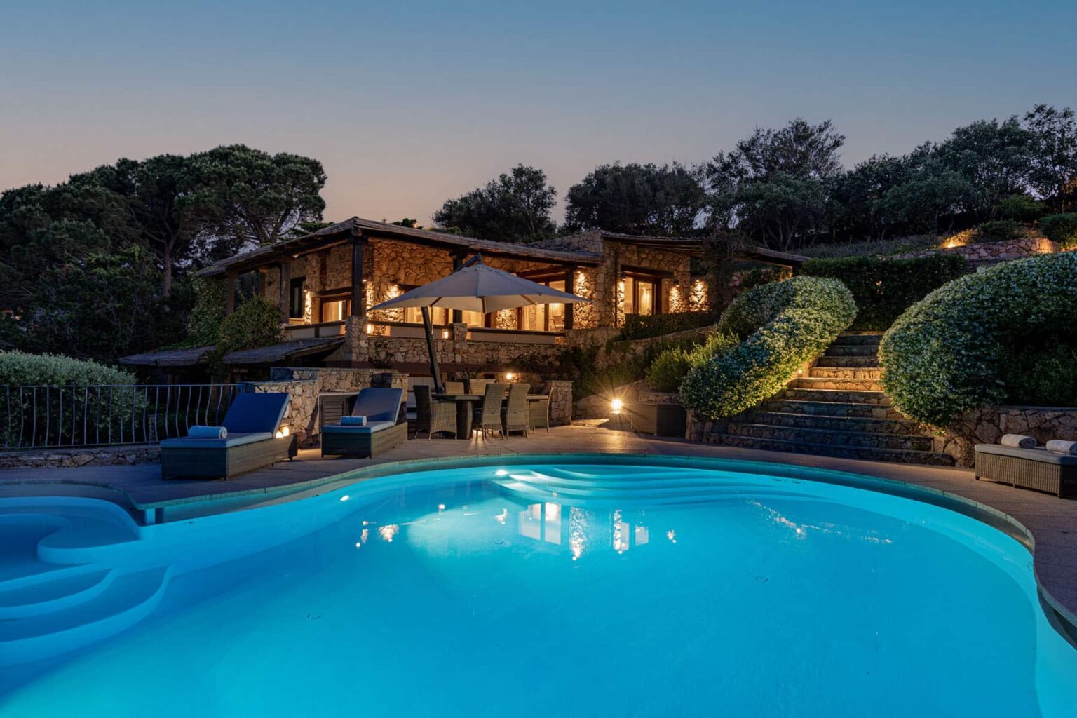 Luxurious outdoor pool illuminated at night, surrounded by landscaped gardens and a modern home with large windows, creating a serene and inviting atmosphere for relaxation and entertainment. | Only on SopranoVillas