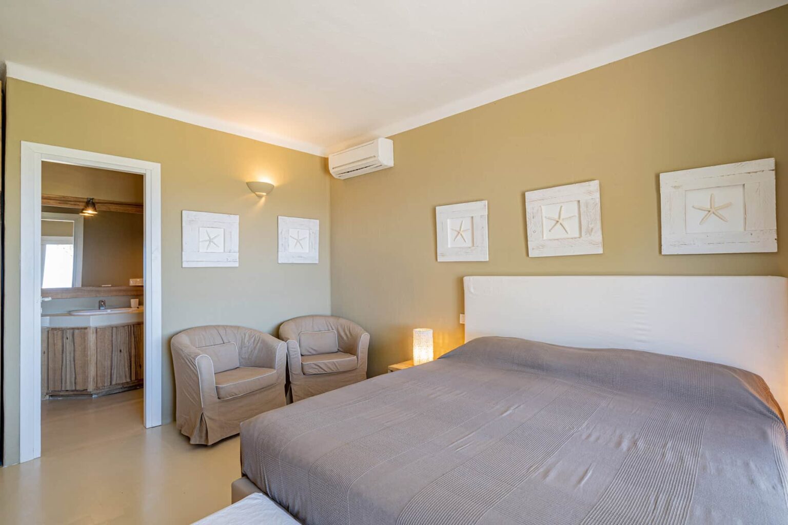 Cozy bedroom with a queen-sized bed, two armchairs, and framed artwork on beige walls, featuring a doorway leading to a bathroom. Natural light illuminates the space, creating a warm and inviting atmosphere. | Only on SopranoVillas