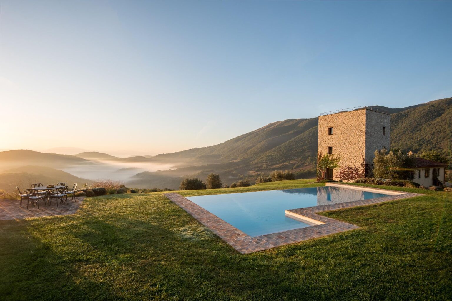 Scenic view of a modern stone villa with a swimming pool, surrounded by lush green hills and misty valleys at sunrise. Outdoor seating area complements the tranquil landscape, ideal for relaxation and enjoying nature. | Only on SopranoVillas