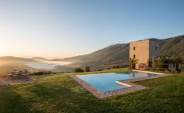 Scenic view of a modern stone villa with a swimming pool, surrounded by lush green hills and misty valleys at sunrise. Outdoor seating area complements the tranquil landscape, ideal for relaxation and enjoying nature.