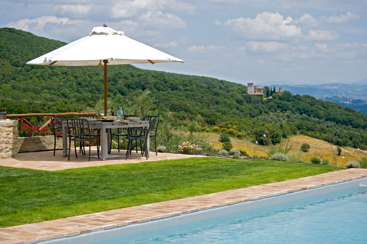 Scenic outdoor dining area with a wooden table and black chairs under a large white umbrella, overlooking lush green hills and a distant castle. The patio features a stone border and a swimming pool, creating a perfect setting for relaxation and enjoyment in nature. | Only on SopranoVillas