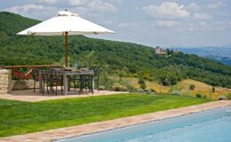 Scenic outdoor dining area with a wooden table and black chairs under a large white umbrella, overlooking lush green hills and a distant castle. The patio features a stone border and a swimming pool, creating a perfect setting for relaxation and enjoyment in nature.