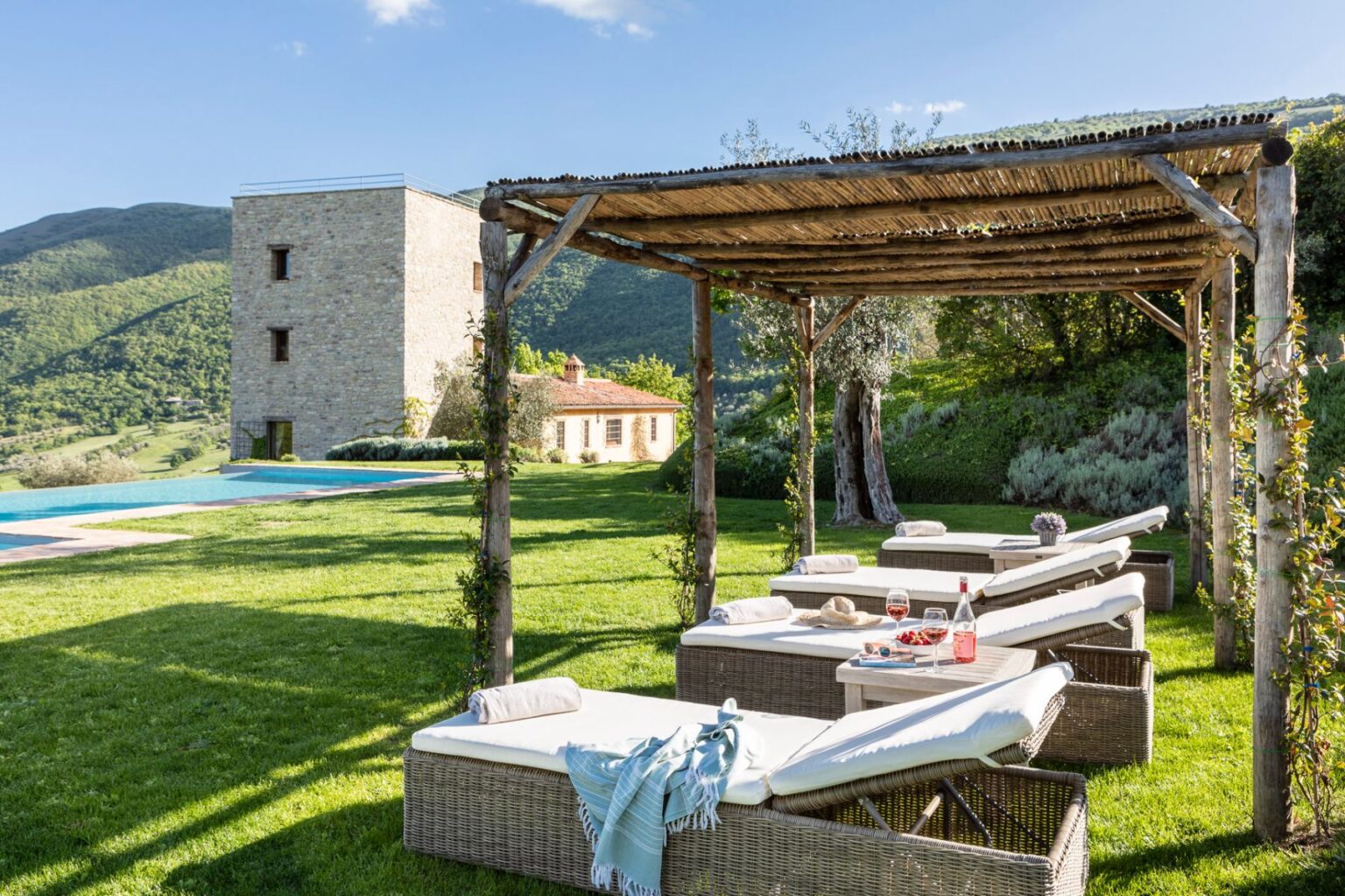 Luxurious outdoor lounge area with sunbeds under a wooden pergola, featuring a table set with wine and snacks, overlooking a scenic landscape with rolling hills and a swimming pool in the background. Ideal for relaxation and enjoying nature. | Only on SopranoVillas