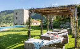 Luxurious outdoor lounge area with sunbeds under a wooden pergola, featuring a table set with wine and snacks, overlooking a scenic landscape with rolling hills and a swimming pool in the background. Ideal for relaxation and enjoying nature.