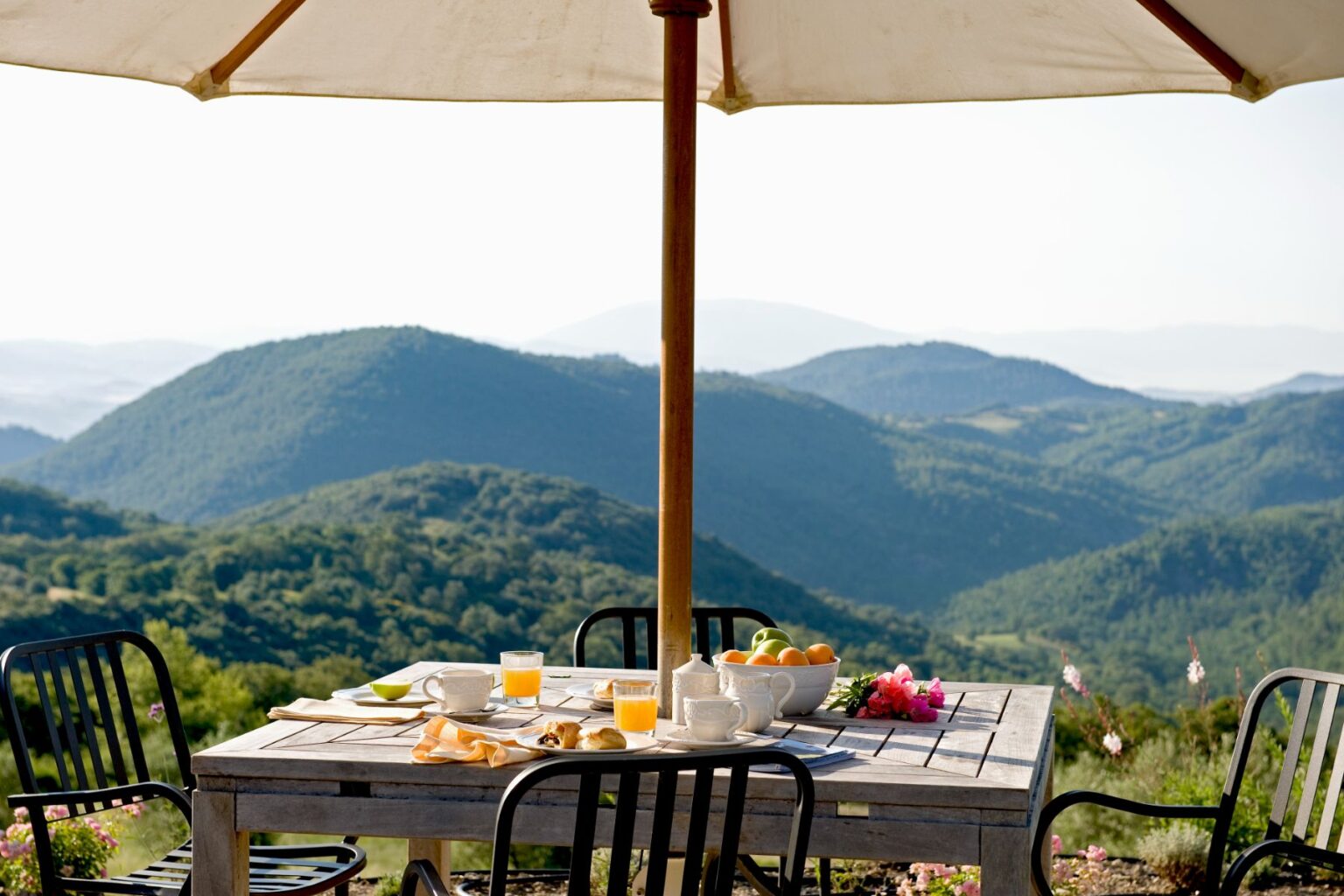 Outdoor breakfast setting with a wooden table under a large umbrella, featuring fresh fruit, pastries, and drinks, overlooking lush green mountains and a clear sky. Perfect for a serene morning getaway. | Only on SopranoVillas