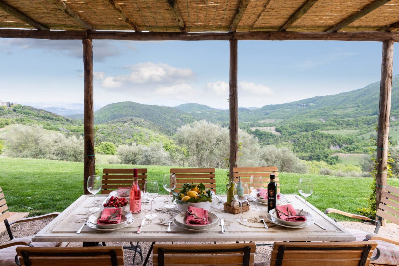 Outdoor dining setup with a scenic view of rolling green hills, featuring a table set with plates, wine glasses, a bottle of rosé, and a bowl of fresh fruit, under a wooden pergola. | Only on SopranoVillas