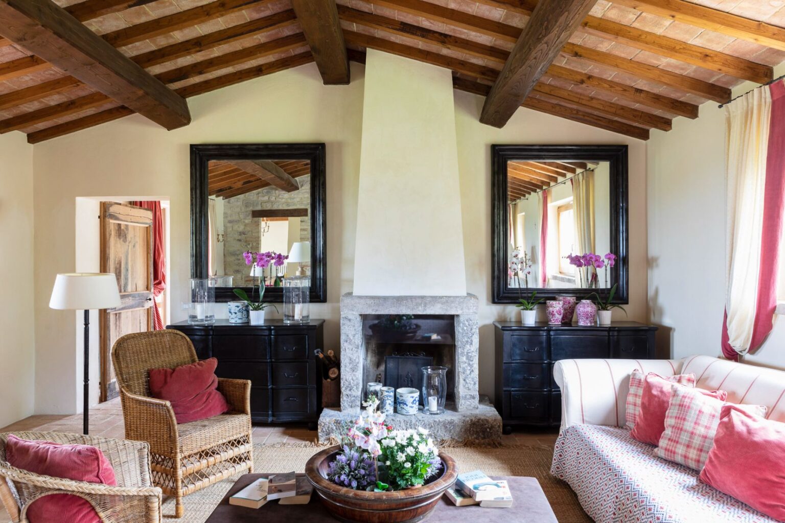 Cozy living room featuring rustic wooden beams, a stone fireplace, and elegant decor. The space includes wicker chairs, a plush sofa with red cushions, and decorative mirrors. Fresh flowers and books add a charming touch to the inviting atmosphere. | Only on SopranoVillas