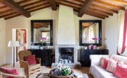 Cozy living room featuring rustic wooden beams, a stone fireplace, and elegant decor. The space includes wicker chairs, a plush sofa with red cushions, and decorative mirrors. Fresh flowers and books add a charming touch to the inviting atmosphere.