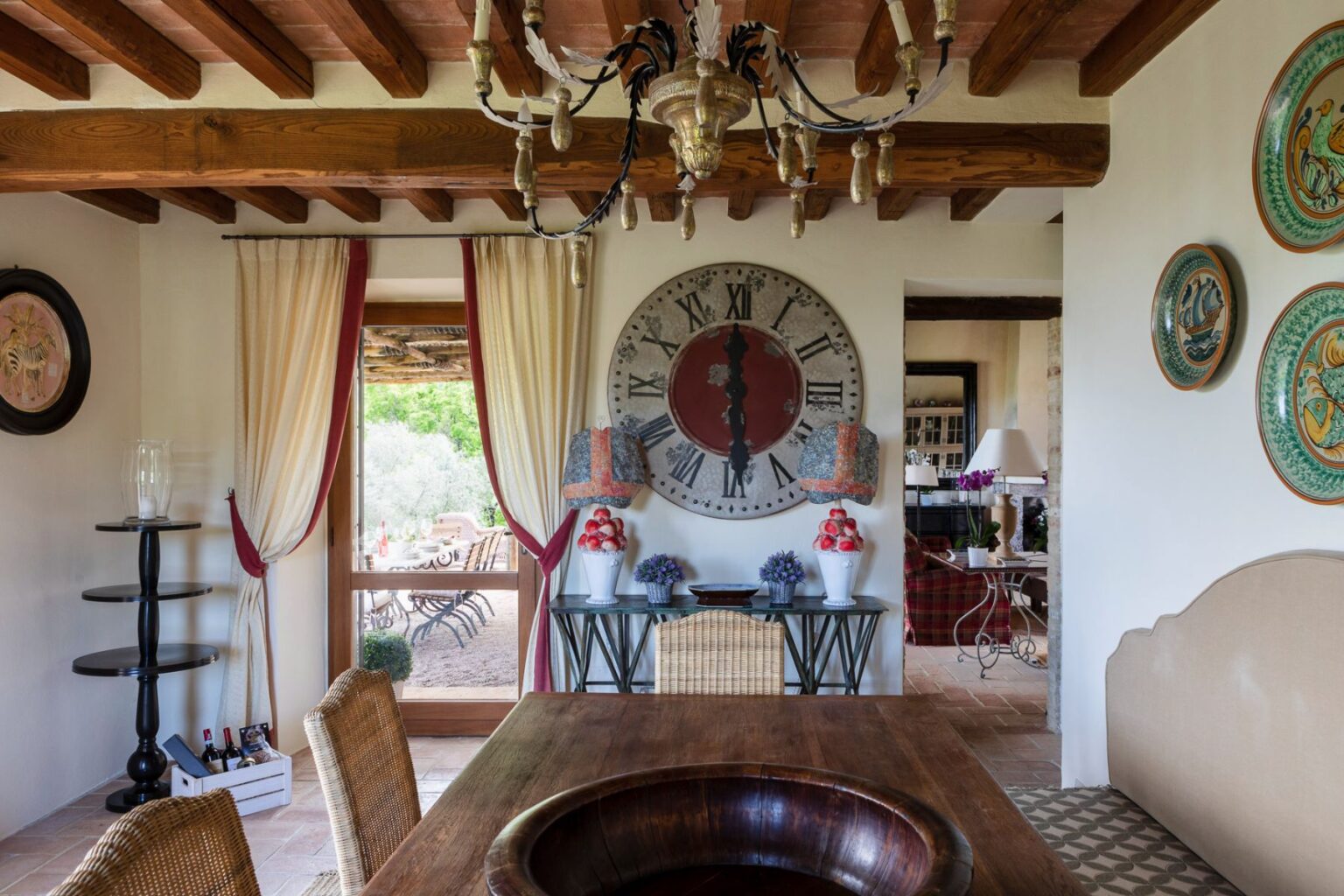 Stylish interior of a cozy dining area featuring wooden beams, a large wall clock, decorative lamps, and a view of an outdoor patio with seating. The room showcases a blend of rustic and elegant decor, highlighted by a round wooden table and colorful wall plates. | Only on SopranoVillas