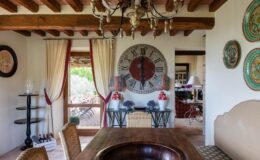 Stylish interior of a cozy dining area featuring wooden beams, a large wall clock, decorative lamps, and a view of an outdoor patio with seating. The room showcases a blend of rustic and elegant decor, highlighted by a round wooden table and colorful wall plates.