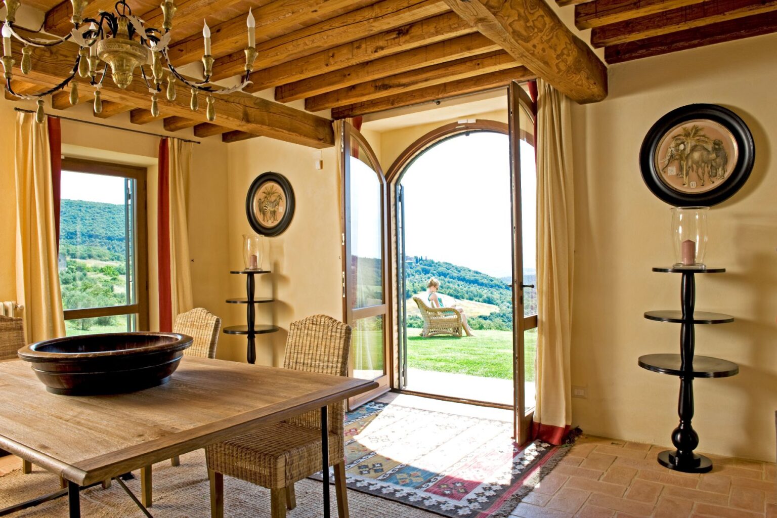 Cozy interior of a rustic dining area featuring a wooden table, woven chairs, and a chandelier, with an open door leading to a scenic outdoor view of lush hills. Natural light floods the space, enhancing the warm tones of the decor. | Only on SopranoVillas