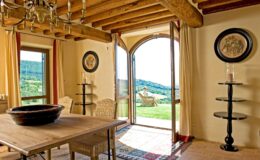 Cozy interior of a rustic dining area featuring a wooden table, woven chairs, and a chandelier, with an open door leading to a scenic outdoor view of lush hills. Natural light floods the space, enhancing the warm tones of the decor.