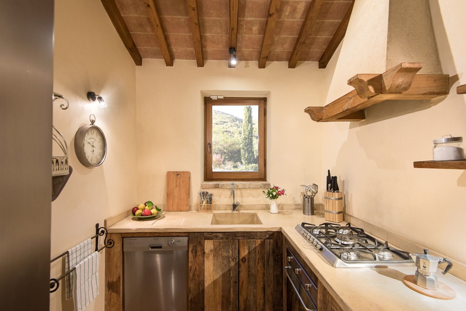 Cozy kitchen interior featuring wooden beams, a window with a scenic view, a stainless steel dishwasher, a gas stove, and decorative elements like a clock and fresh flowers. | Only on SopranoVillas